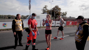 Ulica, kobieta w stroju sportowym pije wodę. Obok niej mężczyzna z obsługi maratonu.