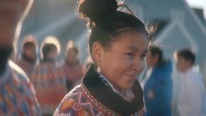 Kadr z filmu: portret młodej kobiety o azjatyckich rysach. Ciemne włosy ma upięte w kok. Ma na sobie kolorową, wzorzystą szatę z wysokim, czarnym kołnierzem. Kobieta stoi na dworze, w otoczeniu współplemieńców.