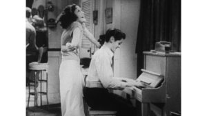 Mężczyzna gra na pianinie, kobieta oparta o jego ramię, śpiewa.. Śmieją się. Kadr czarno-biały.