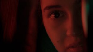 Kadr z filmu: Półmrok, z dominantą czerwieni. Zbliżenie na twarz dziewczyny. Widzimy tylko jedno jej oko, nos, pieprzyk na policzku i lekko rozchylone usta. Reszta rozpływa się w cieniu.