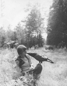 Kadr. Młoda kobieta z dwoma warkoczami dotyka ustami dzioba ptaka, siedzącego na jej kolanie. W ręce trzyma aparat fotograficzny.