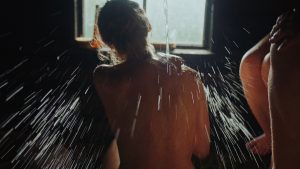 Wnętrze sauny. W półmroku, na tle okna, tyłem do nas siedzi naga kobieta. Ma gęste, spięte w kok, jasne włosy. Na jej plecy spływa z góry strumień wody, rozpryskując się wkoło.