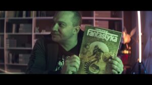 Kadr z filmu. Portret pisarza, Łukasza Orbitowskiego - krótko ostrzyżony mężczyzna w ciemnej bluzie. Siedzi na tle regałów z książkami. Do kamery pokazuje archiwalny numer miesięcznika „Fantastyka”.