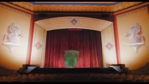 Wnętrze starego teatru. Widok na okno sceniczne zasłonięte czerwoną kurtyną. Widownia jest pusta. Na środku sceny, przed kurtyną unosi się obłok zielonego dymu.