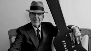 Kadr z filmu. Piosenkarz Leonard Cohen siedzi, obejmując gitarę w czarnym futerale. Ma na sobie koszulę i marynarkę w paski, krawat, szary kapelusz.