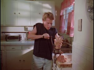 Kadr z filmu. Młody mężczyzna w kuchni. Ma na sobie czarną koszulkę i jasne dżinsy. Wkłada odciętą ludzką dłoń do maszynki do mięsa, z której wylatuje mielone mięso.