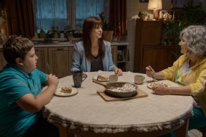 Kadr z filmu. Przy okrągłym stole w kuchni siedzą trzy osoby: chłopiec z nadwagą w niebieskiej koszulce, kobieta w średnim wieku, z długimi włosami, w błękitnej marynarce, starsza kobieta z siwymi, kręconymi włosami, w żółtej bluzie. Przed nimi stoją talerze z posiłkiem, na środku stołu patelnia z tym samym daniem.