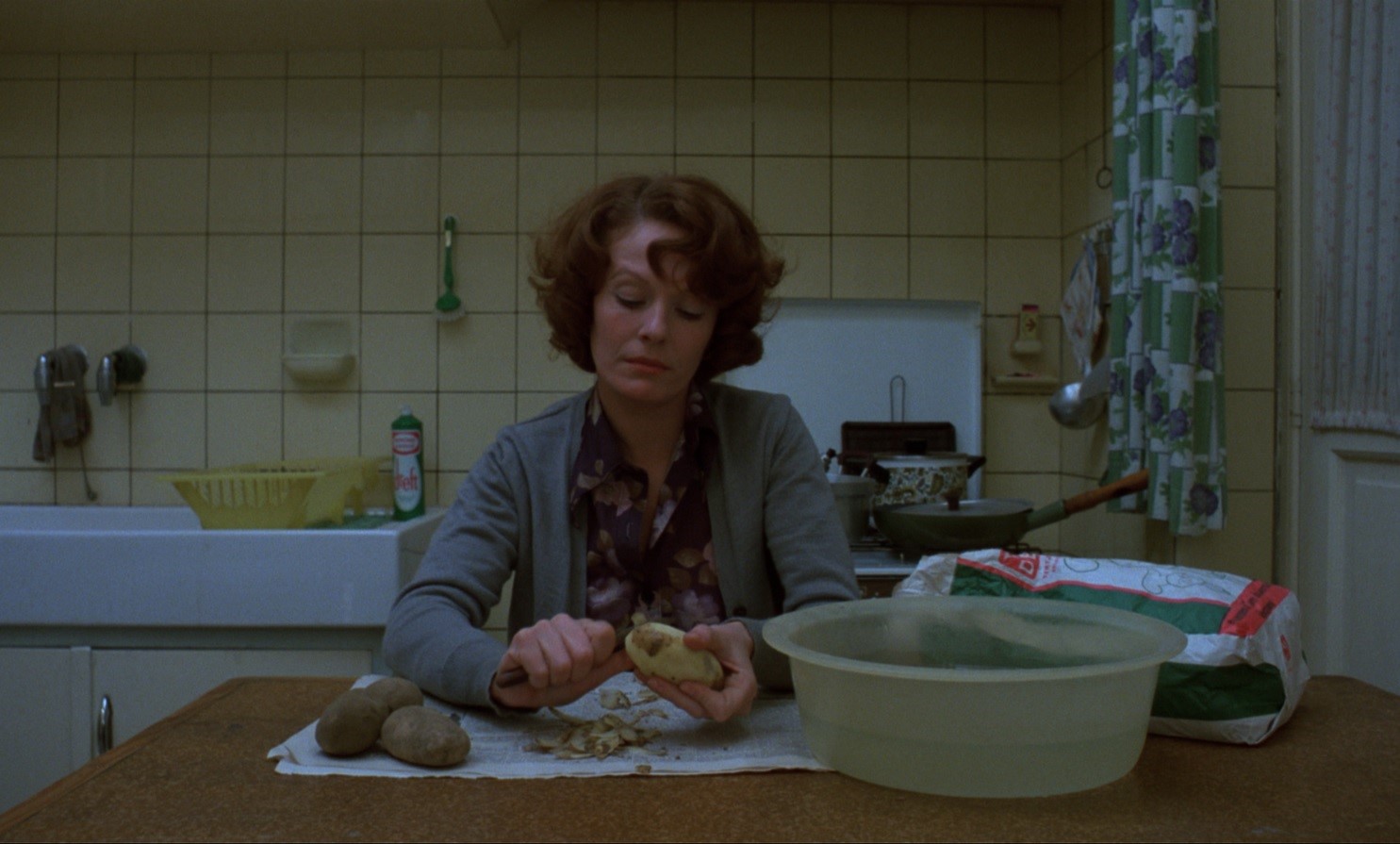 Kadr z filmu. Kobieta w kwiecistej bluzce, szarym sweterku, w kuchni. Siedzi przy stole i obiera ziemniaki.