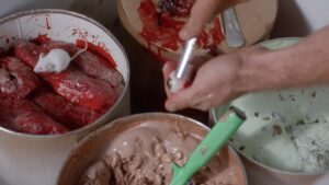 Kadr z filmu. Trzy naczynia wypełnione masami - lodowymi i zakrwawionym mięsem. Po mięsie biega biała mysz. Na pierwszy planie widać dłonie trzymające ludzką gałkę oczną.