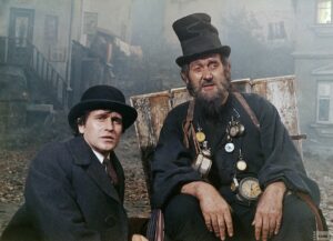 Kadr z filmu. Dwóch mężczyzn ubranych na czarno, w czarnych nakryciach głowy. Jeden z nich jest obwieszony zegranami o różnych rozmiarach. W tle zamglone podwórze i budynki mieszkalne.