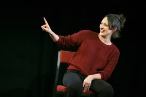 Kadr z filmu. Kobieta na czerwonym krześle, w ciemnoczerwonym swetrze i czarnych spodniach, uśmiecha się i unosi dłoń z wysuniętym wskazującym palcem w prawą stronę.