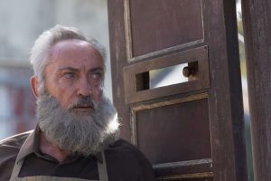 Mężczyzna z siwymi włosami i siwą brodą stoi przy otwartych drzwiach. Kadr z filmu.