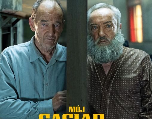 Plakat promujący film "Mój sąsiad Adolf". Dwóch starszych mężczyzn - jeden łysiejący, w niebieskiej koszulki, drugi - w brązowej piżamie, z siwymi włosami i siwą brodą. Stoją we wnętrzach swoich mieszkań przy ścianie widocznej między nimi (w przekroju).