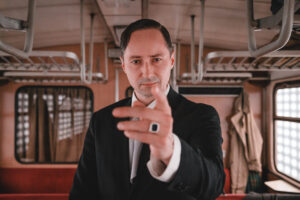 Kadr z filmu. Elegancki mężczyzna w garniturze, z gładko przyczesanymi włosami i sygnetem na palcu, wskazuje w stronę obiektywu. W tle wnętrze pociągu.
