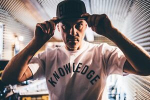 DJ Pestka- młody mężczyzna w białej koszulce z napisem, w czapeczce z daszkiem