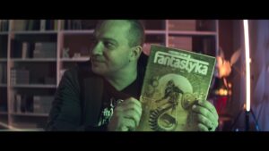 Kadr z filmu. Mężczyzna ubrany na czarno trzyma numer pisma "Fantastyka". W tle półki z książkami i figurkami.