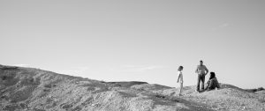 Kadr z filmu LIMBO, czarno-biały. Na skalistym wzgórzu stoi dziewczynka, mężczyzna, obok siedzi Aborygenka. Postaci widzimy z oddali, po prawej stronie kadru wypełnionego krajobrazem.