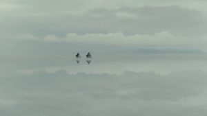 Kadr z filmu. Widziane z oddali dwie postaci na rowerach. W ich otoczeniu i tle widać chmury i wodę.