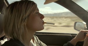 Młoda kobieta z blond włosami i papierosem w ustach prowadzi samochód. Widzimy ją z profilu. W tle, za oknem auta, plener.