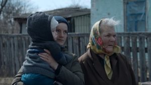 Kadr z filmu. Na tle budynków i drewnianego płotu stoi starsza kobieta w kwiecistej chuście, młodsza kobieta w kurtce i czapce, z dzieckiem na rękach.