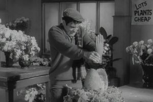 Kadr z czarno-białego filmu. W kwiaciarni, mężczyzna z zawziętą miną trzyma odciętą ludzką dłoń nad bryłą wyglądającą nieco na roślinę, nieco na jajo.