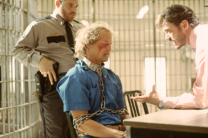 kadr z filmu. W więzieniu, przy stole siedzi zakuty w kajdany mężczyzna z półdługimi blond włosami i raną na twarzy. Na wprost rozmówca w jasnej koszulki, gestykuluje ręką. Za siedzącym funkcjonariusz policji