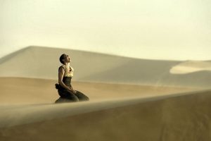 Kadr z filmu. Kobieta w krótkich włosach, koszulce, spodniach siedzi na ziemi w surrealistycznie wyglądającym pustynnym otoczeniu, krzyczy, odchylając głowę w tył.