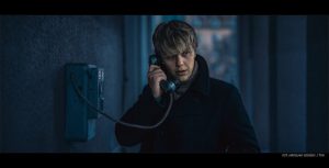 kadr z filmu. młody mężczyzna w blond włosach, niedbale opadających na czoło, w czarnym ubraniu, rozmawia przez telefon stacjonarny.