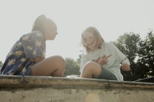 Kadr z filmu. Dwie dziewczyny kolorowo ubrane siedzą na murku, śmieją się i rozmawiają.