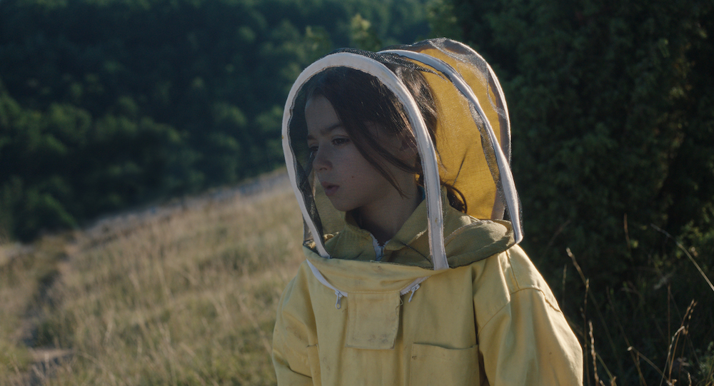 Kadr z filmu. Dziewczynka w stroju pszczelarskim, w kapeluszu pszczelarskim z siatką na twarzy, w kolorze bladożółtym. W tle widać pole i krzaki.