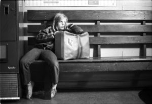 Czarno-biały kadr. Dziewczynka siedzi niedbale na ławce, oparta o torbę podróżną. Ma blond długie włosy, ubrana jest w ciemne spodnie i ciemną kurtkę.