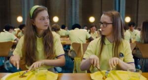 Kadr z filmu. Stołówka. Dwie nastolatki z długimi włosami trzymają sztućce i kroją leżący na ich talerzach kawałek jedzenia. Są ubrane w jasnozielone koszulki, podobnie jak inne osoby w tym samym wieku, widoczne przy stołach w tle.