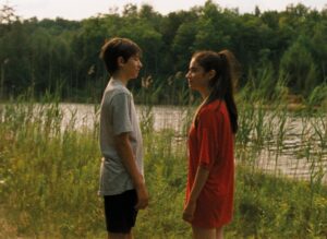 Kadr z filmu. Kilkunastoletni chłopak w szarym t-shircie i ciemnych spodenkach i nastolatka z długimi, związanymi w kitkę włosami, w czerwonym długim t-shircie, stoją na przeciw siebie na tle jeziora.