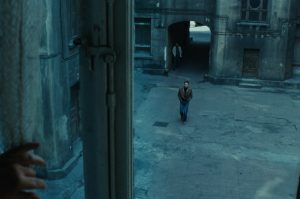 Kadr z filmu. Widoczny przez okno na piętrze mężczyzna idący przez podwórze kamienicy. Widać framugę okna i dłoń w jej pobliżu.