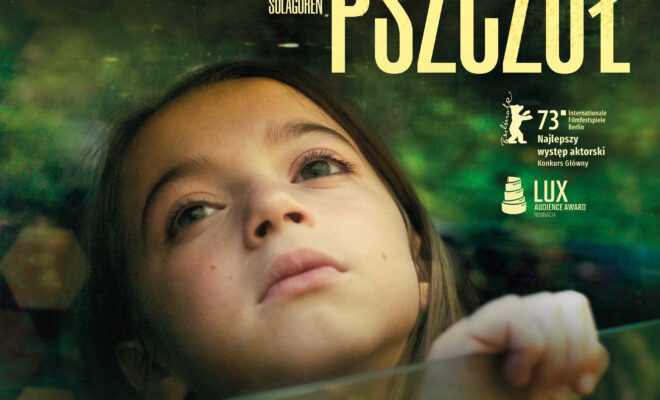 Dziewczynka leży, w tle zieleń. Patrzy w górę, trzymając szybę przed klatką piersiową.