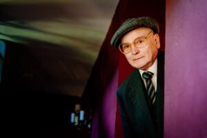 Starszy mężczyzna w okularach, w kaszkiecie, marynarce, krawacie w paski. Wychyla się zza framugi drzwi osadzonych we fioletowej ścianie.