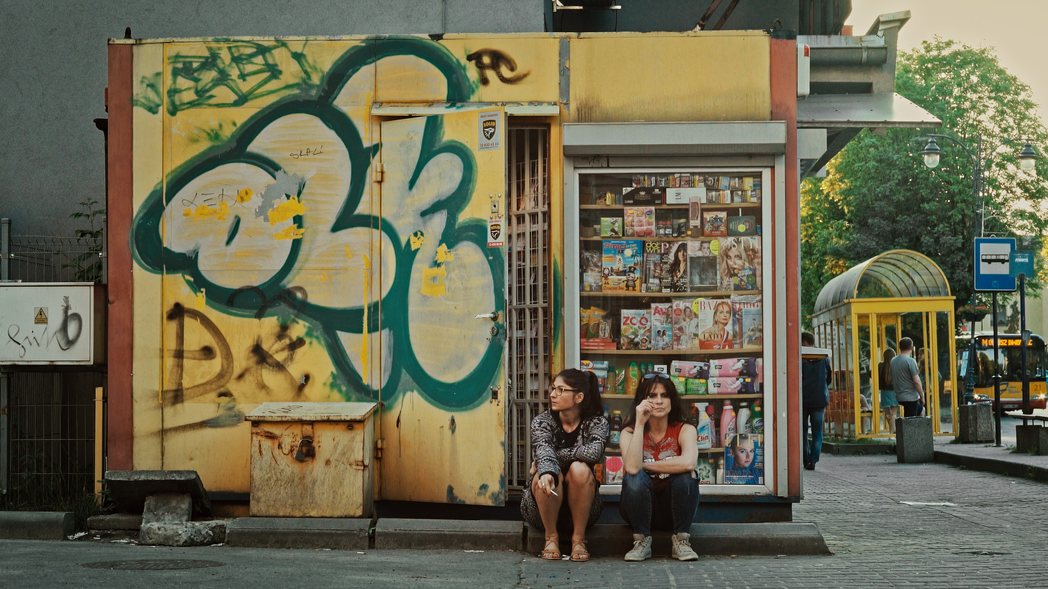 Kiosk pomalowany sprayami, w szybie widać porozkładane magazyny i inne produkty, na schodku siedzą dwie kobiety, palą papierosy.