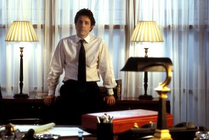 Mężczyzna w białej koszuli, krawacie, spodniach od garnituru, stoi oparty o mebel. Wokół widać wnętrze biura lub salonu, z dwiema nocnymi lampkami zapalonymi.