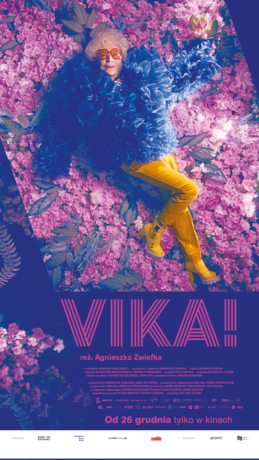 Plakat. Starsza kobieta, dj Vika, leży w żółtych spodniach i pierzastej niebieskiej kurtce na płatkach kwiatów.