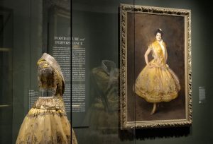 suknia żółta balowa na manekinie, w tle obraz z podobną suknią. Ubrana jest w nią modelka.