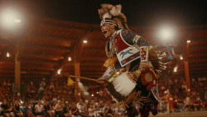 Stadion z drewnianymi trybunami, wypełnionymi widzami. Na pierwszym planie, taniec rytualny wykonuje przedstawiciel rdzennego, indiańskiego plemienia. Ma na sobie tradycyjny strój, na głowie nosi pióropusz.