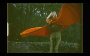 Kadr z archiwalnego filmu ukazuje młodą kobietę, uśmiechniętą blondynkę stojąca na tle lasu. Ubrana w spodnie i ciemną bluzkę ma na sobie intensywnie pomarańczową pelerynę, którą dynamicznym gestem unosi do góry.