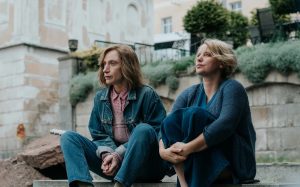 dwie kobiety z jasnymi długimi włosami, w niebieskich codziennych ubraniach, siedzą na murku w mieście