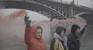 Trzy kobiety w puchowych, zimowych kurtkach, z kapturami zarzuconymi na głowy idą nabrzeżem rzeki. Dwie z nich w uniesionych wysoko rękach niosą dymiące race. W tel widać stalowe przęsła mostu.