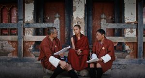 Na schodach prowadzących na taras starego domu siedzi trzech mężczyzn, rozmawiających ze sobą. Mają na sobie tradycyjny bhutański strój. W środku siedzi młodych buddyjski mnich. Mężczyzna z lewej oraz drugi, z prawej na kolanach przed sobą trzymają, spięte, białe kartki.