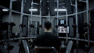 W laboratorium, tyłem do widza, na krześle siedzi mężczyzna. Otacza go aluminiowy stelaż, na którym zamocowane są lampy fotograficzne - tzw. softbox’y oraz liczne aparaty fotograficzne. W głębi na ekranie widać obraz ludzkiej twarzy.