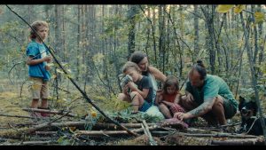 W gęstym lesie, pomiędzy drzewami siedzi skupiona czteroosobowa rodzina: kobieta, mężczyzna i dwoje małych dzieci. Obok nich stoi jeszcze jedno dziecko - chłopiec w niebieskim t-shircie i krótkich spodenkach.