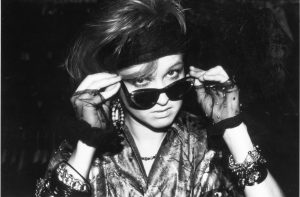 Archiwalne, czarno-białe zdjęcie piosenkarki Cyndi Lauper. Młoda dziewczyna o włosach spiętych opaską, spogląda na nas zza ciemnych okularów opuszczonych na czubek nosa. Na dłoniach trzymających okulary ma koronkowe rękawiczki.