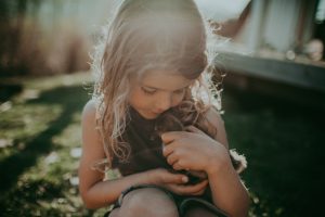 W ogrodzie, oświetlona promieniami słońca, kuca mała dziewczynka. W dłoniach, przy policzku trzyma kacze pisklę.