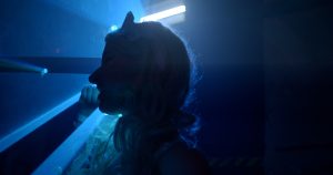 W półmroku, w kontrastowym świetle padającym od fluorescencyjnej lampy widać profil dziewczęcej głowy. Blond loki spina finezyjna kokarda.